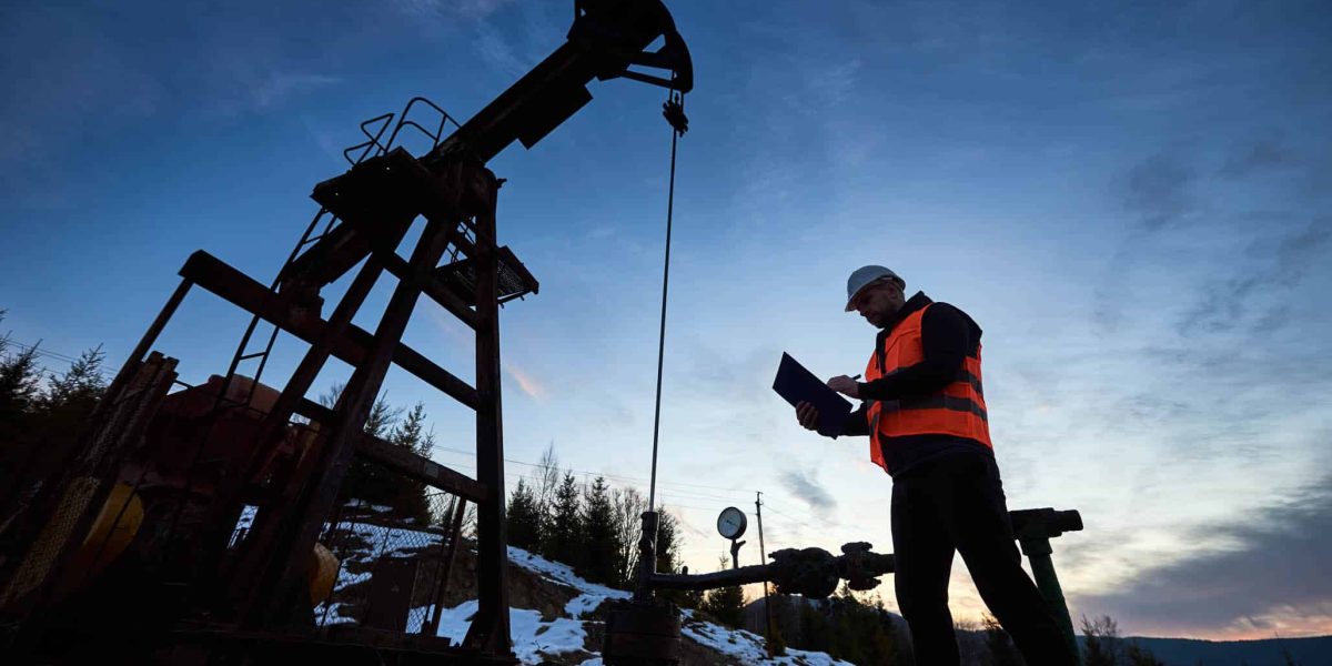 Low angle of petroleum engineer in work vest looking at oil pump rocker-machine and writing on clipboard. Oil worker checking work of balanced beam petroleum pump jack under beautiful evening sky.